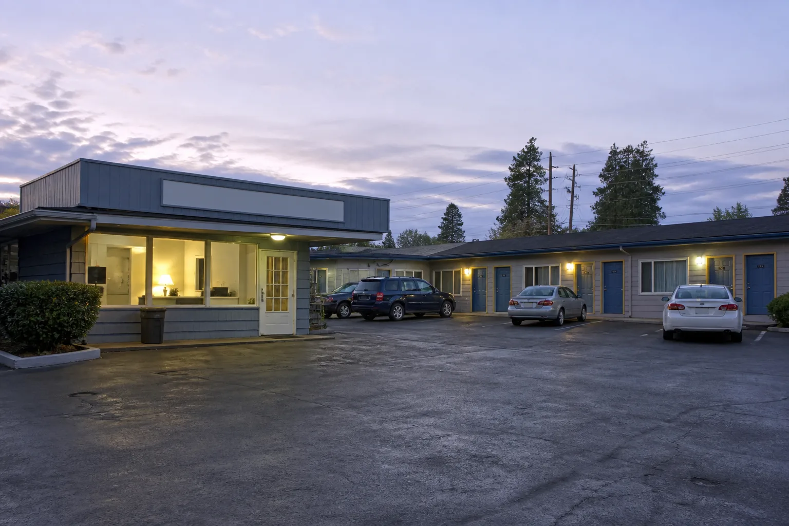 Roadside motel office at dusk with a warm interior lamp, neon vacancy strip unlit, asphalt lot with a few cars, numbered doors stretching to the right, 1600x900