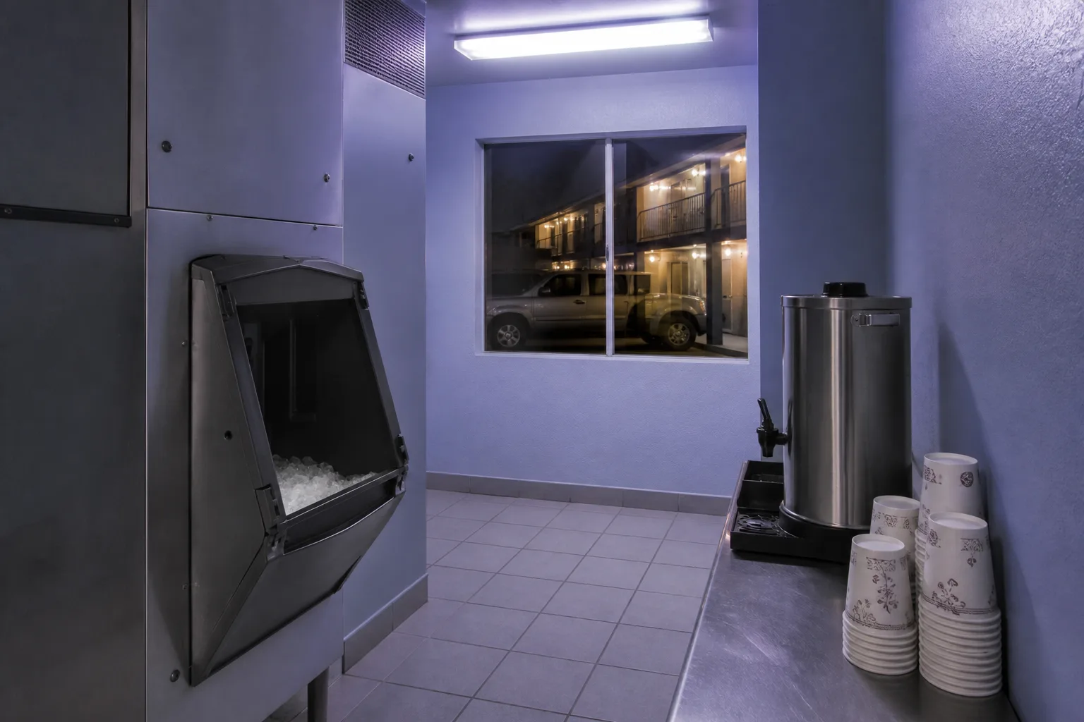 Small motel ice and coffee room at night, metal ice machine door half open, paper cups stacked by a coffee urn, fluorescent light over a narrow tiled floor, 1400x900